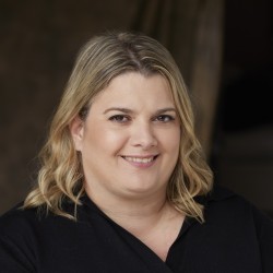 A young blond woman with shoulder length blond hair, a smile, wearing a black v-necked top and a dark background.